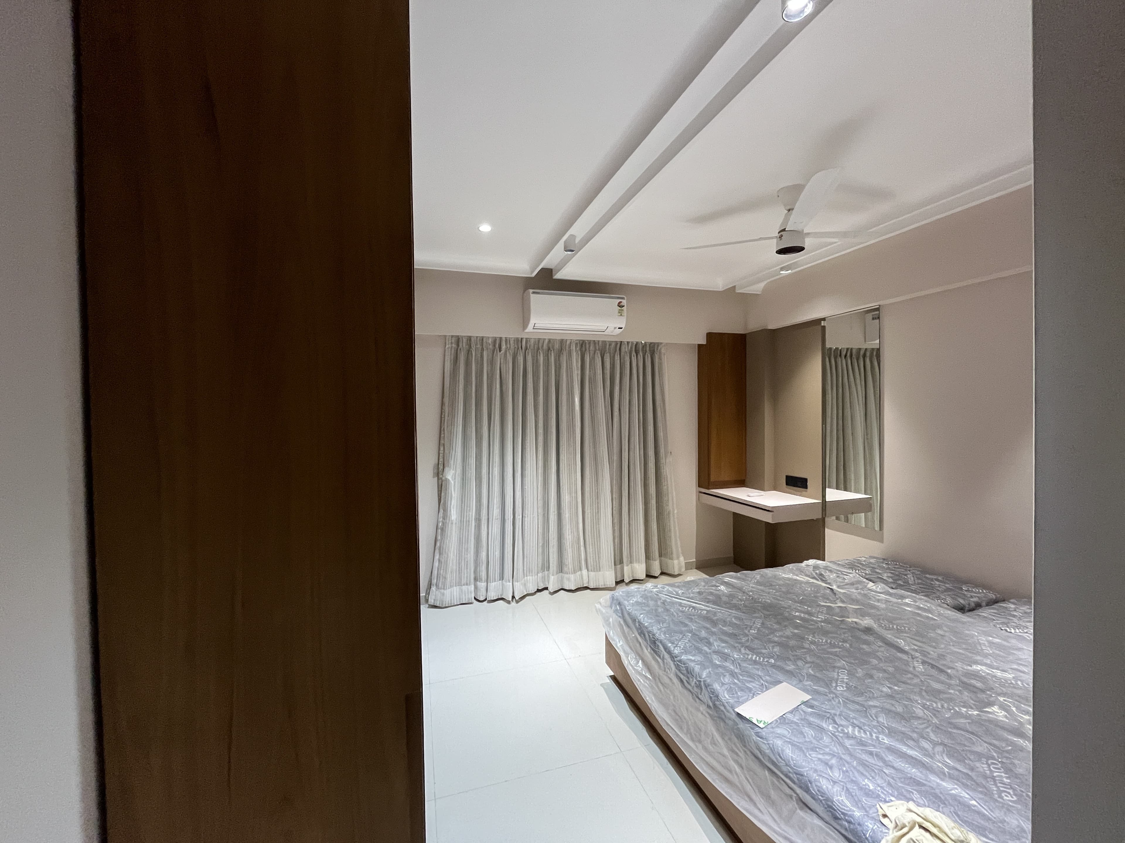 Modern bedroom with wooden wardrobe, sheer curtains, ceiling fan and neutral palette by BR Interiors