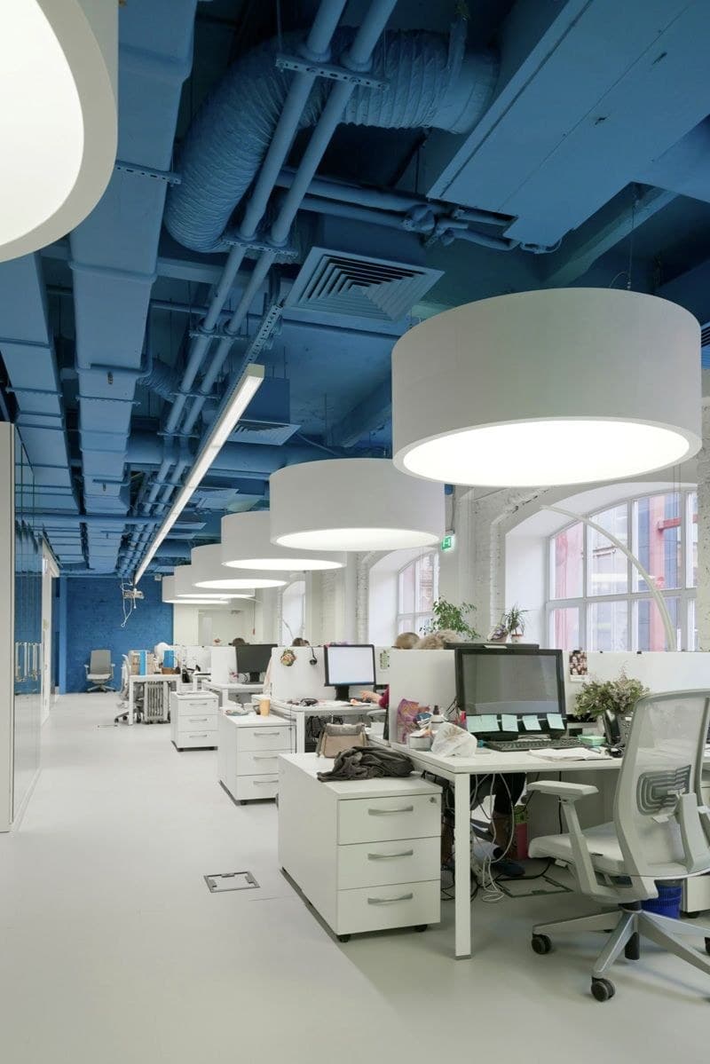 Industrial-modern office with exposed blue ceiling ductwork, large white drum pendant lights and bright white workstations